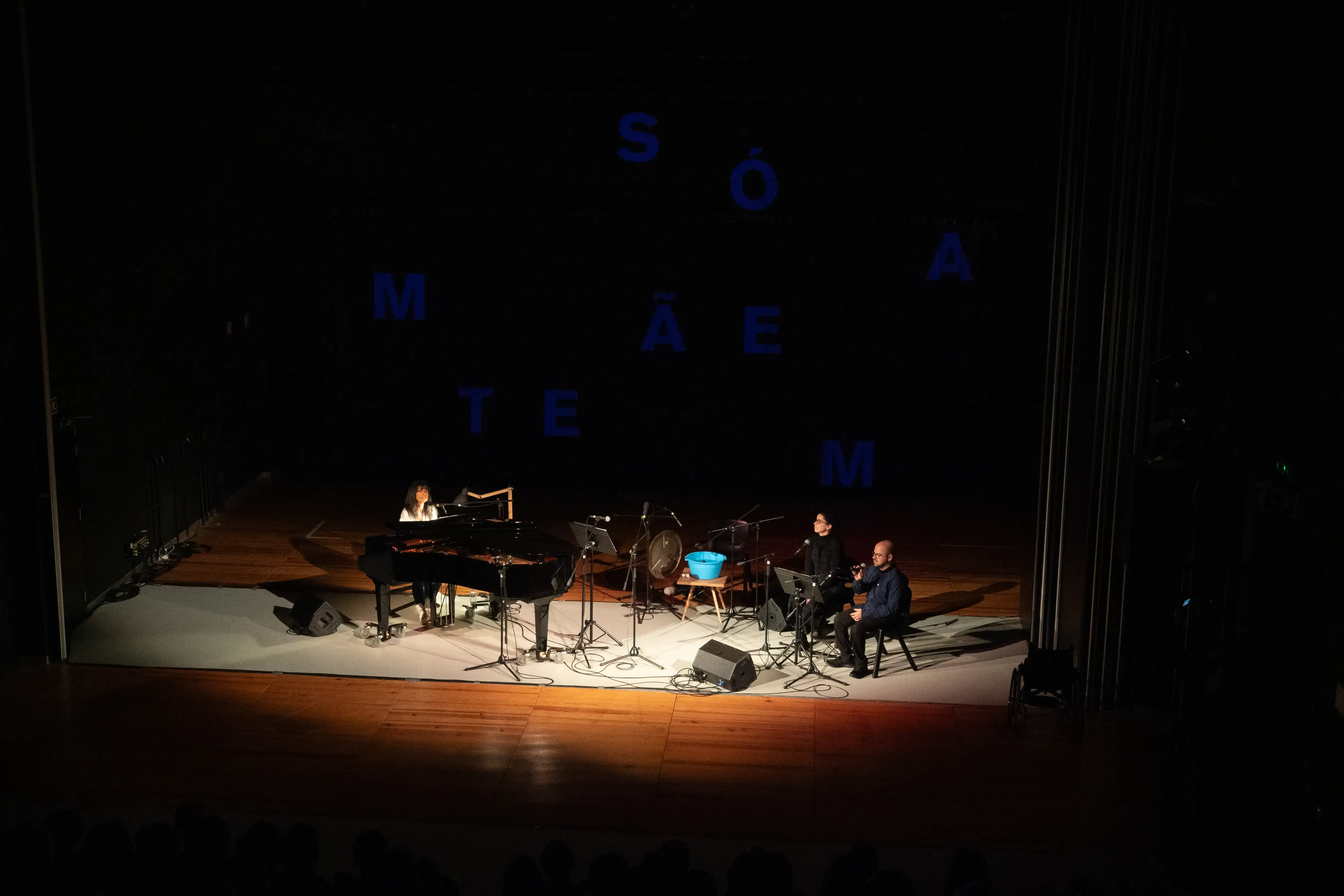 Uma mulher está a tocar piano num palco. No lado direito da imagem duas pessoas acompanha com voz. Ao fundo tem uma projeção de várias letras separadas em azul.